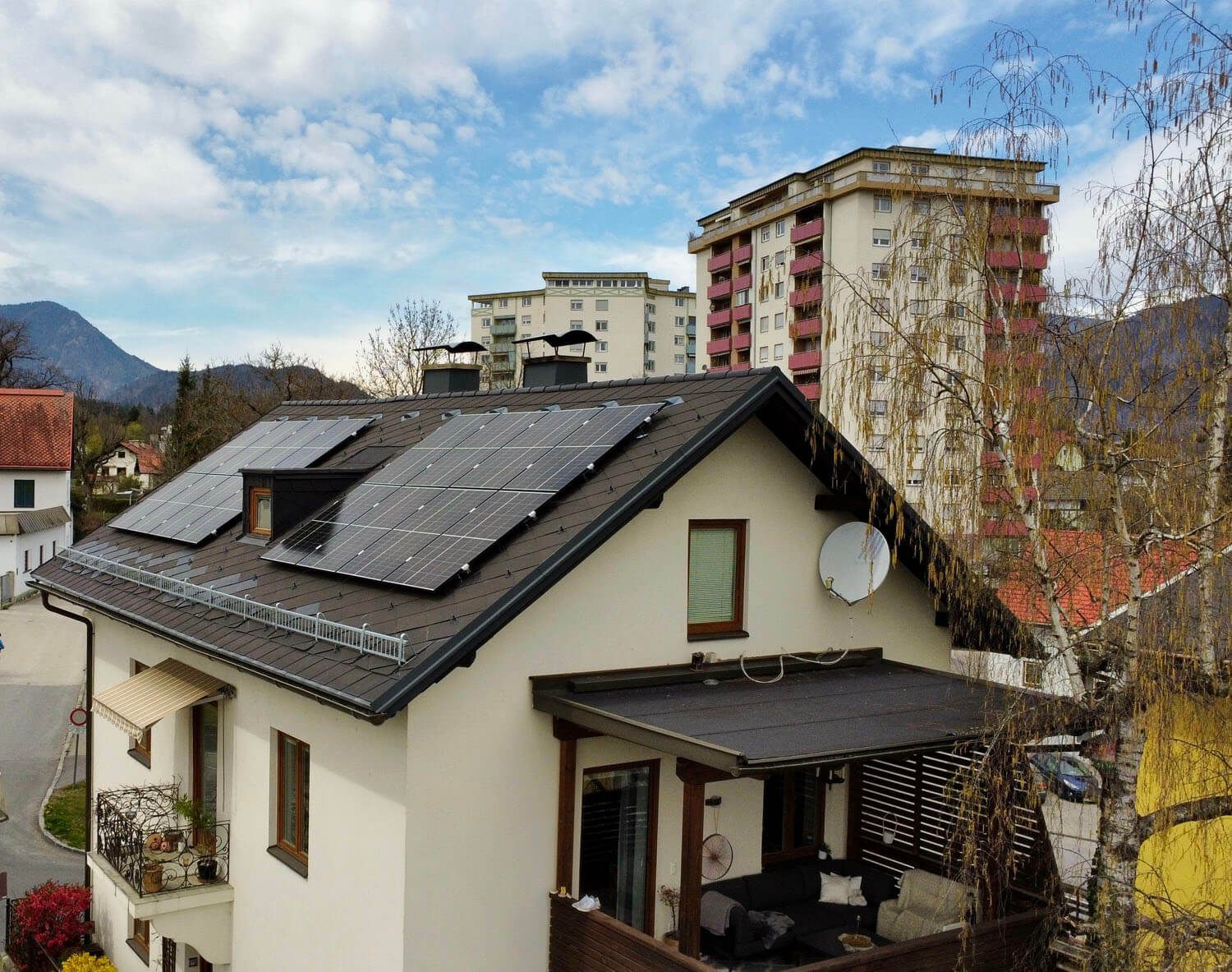 Wohnhaus mit Balkon und dunklem Satteldach mit PV-Modulen, im Hintergrund Wohntürme und Berge.