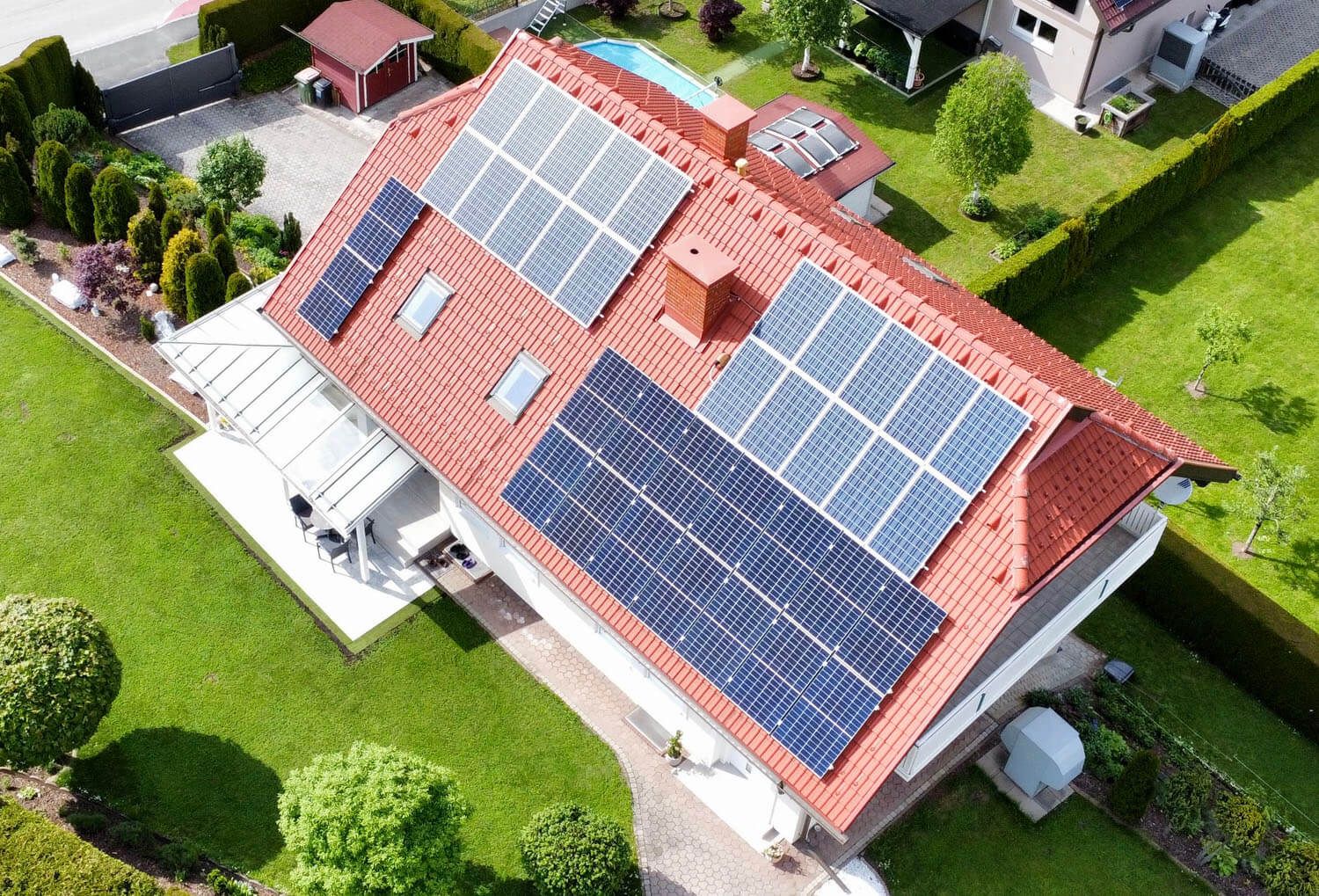 Schrägluftbild eines Hauses mit rotem Ziegeldach und PV-Modulen auf zwei Dachflächen, im Garten ein Pool.