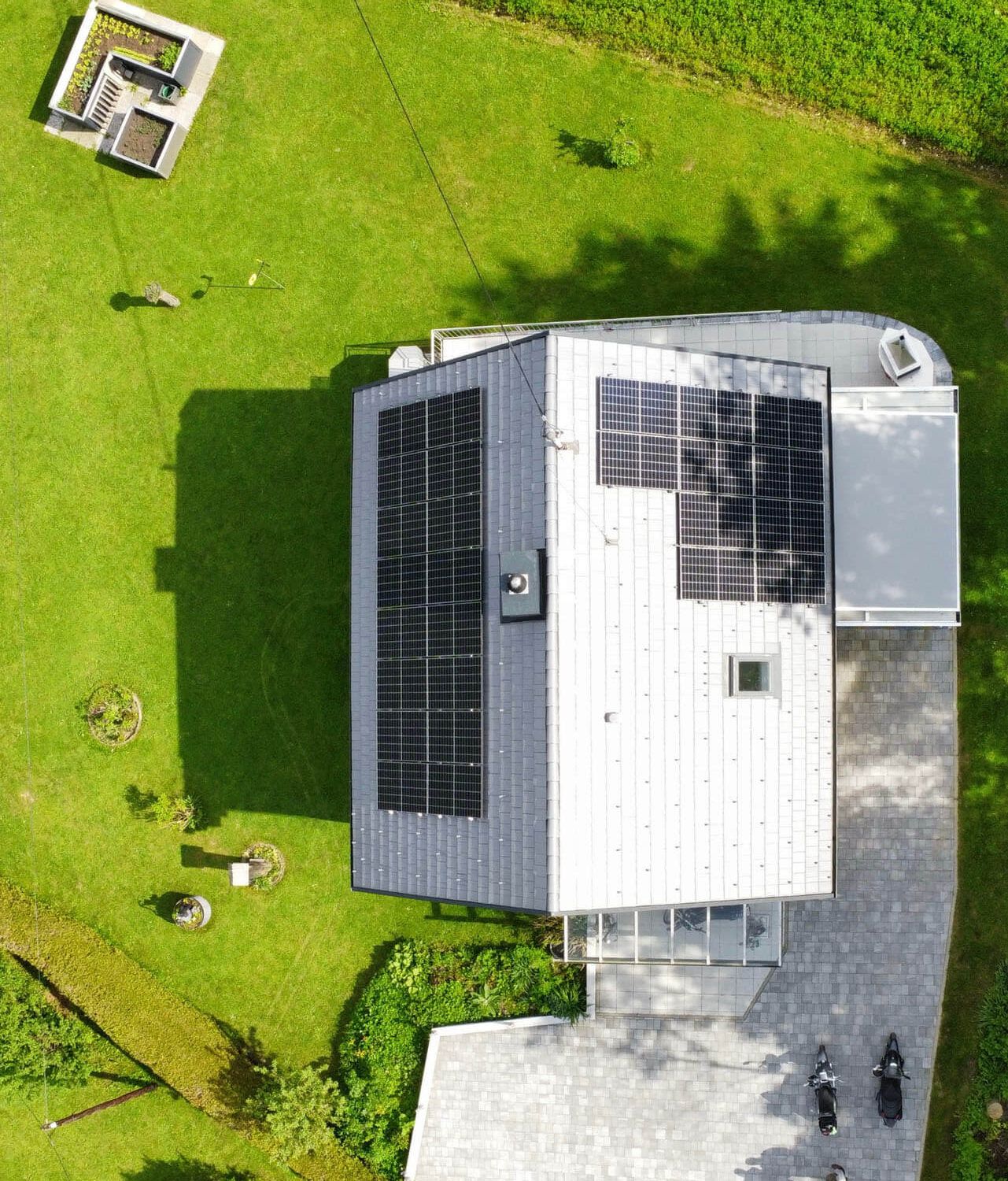 Senkrechte Draufsicht auf ein Haus mit hellem und dunklem Dachteil, beide mit PV-Modulen belegt, davor gepflasterte Einfahrt und Rasen.