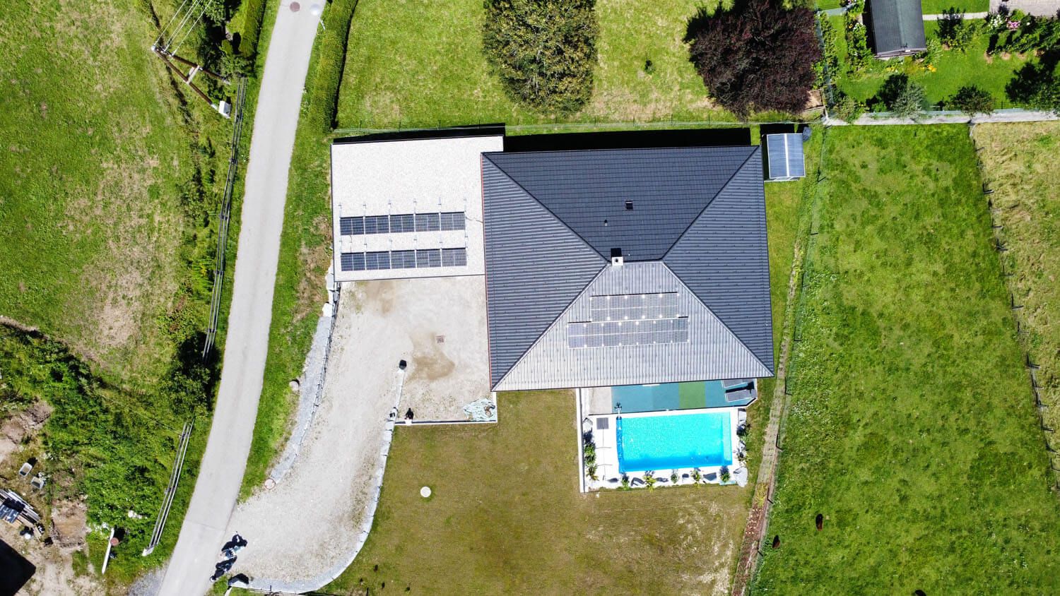 Draufsicht auf ein Haus mit grauem Walmdach und PV-Modulen auf Dach und Flachdach-Zubau, davor ein Pool.