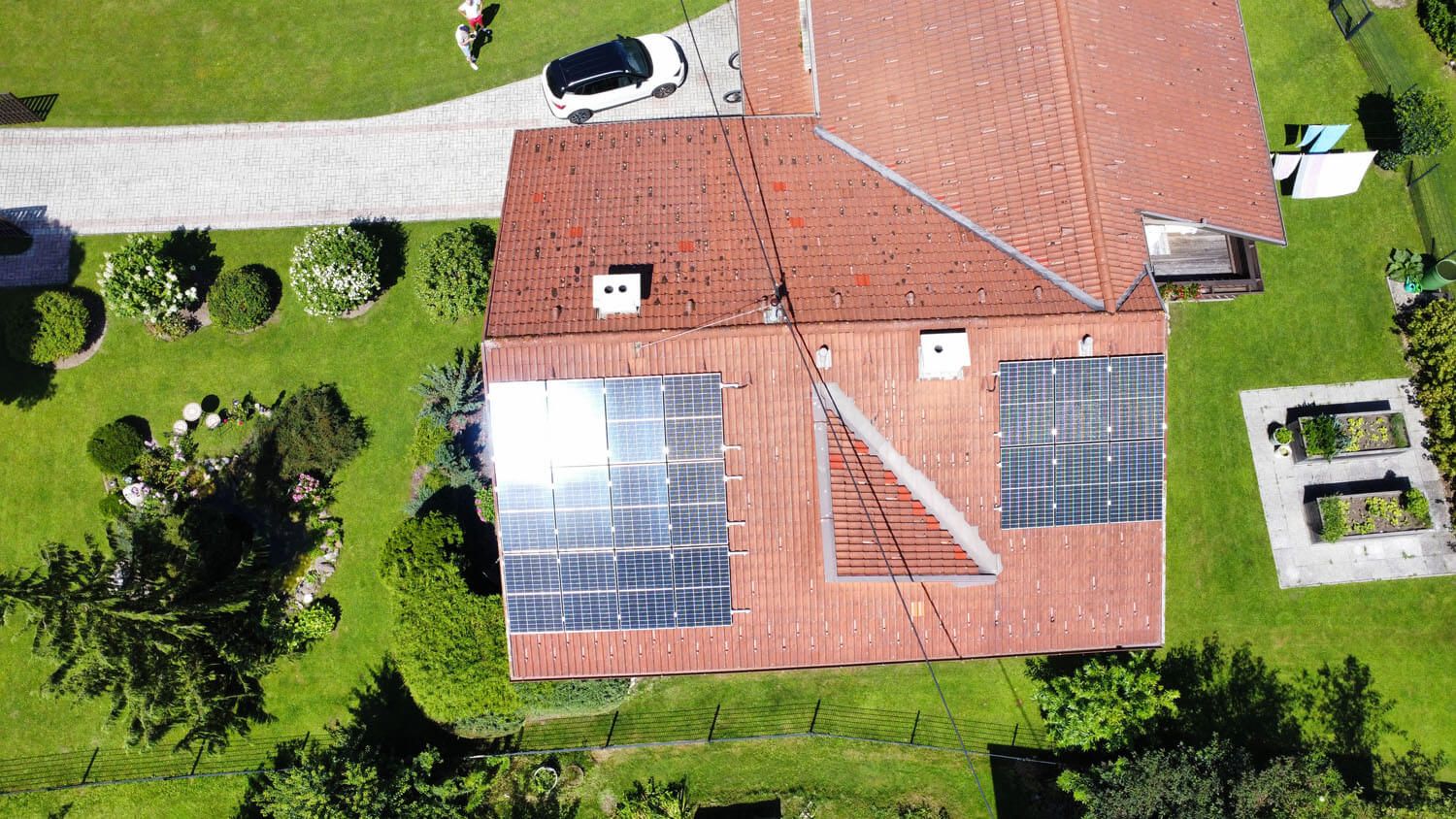 Einfamilienhaus mit rotem Ziegeldach und PV-Modulen auf zwei Dachseiten, umgeben von Garten.