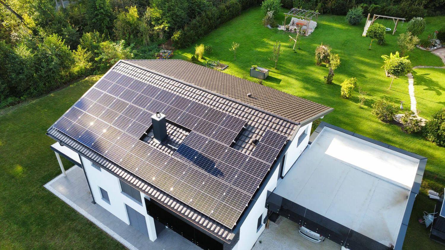 Luftaufnahme eines weißen Einfamilienhauses, dessen dunkle Dachfläche vollständig mit PV-Modulen belegt ist; daneben ein Flachdach-Zubau und eine große Wiese.